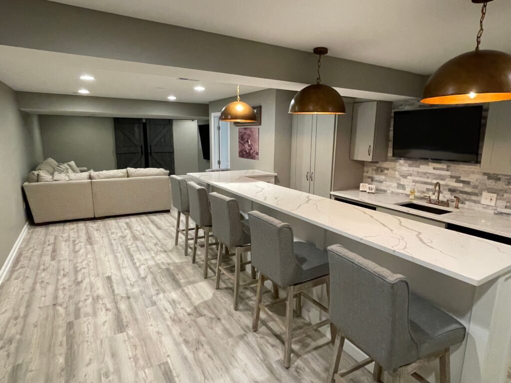 Pittsburgh Basement Finishing project featuring a modern open-concept layout with a spacious white island, bar seating, recessed lighting, and a cozy lounge area with a sectional sofa and wall-mounted TV.
