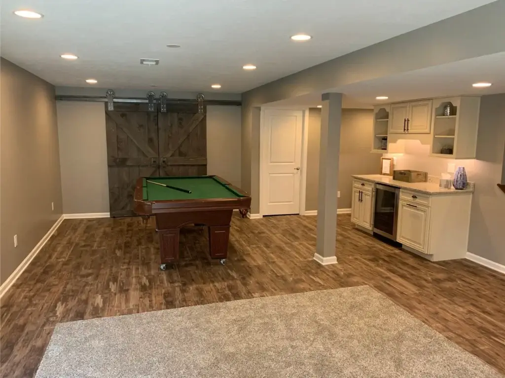 Finished basement remodeling company project built by Pittsburgh Basement Finishing featuring a game room with pool table, rustic barn doors, and a custom wet bar with white cabinetry and granite countertops.